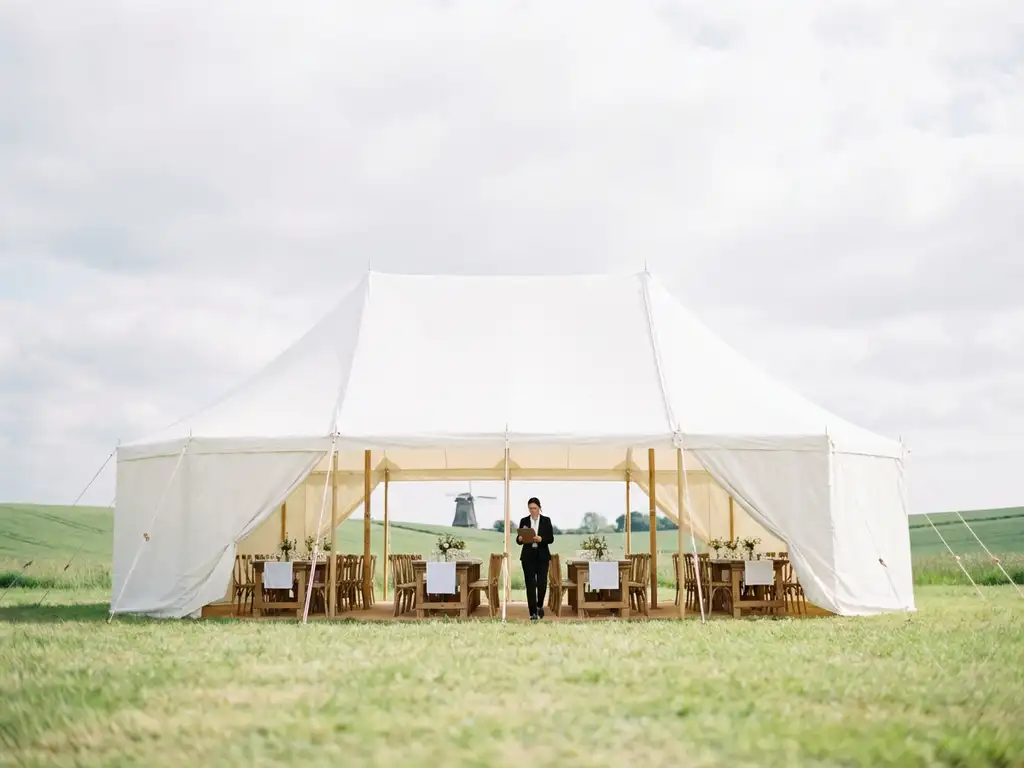 White event tent in Dutch countryside with wooden tables and linens, person preparing for event, windmill visible in distance