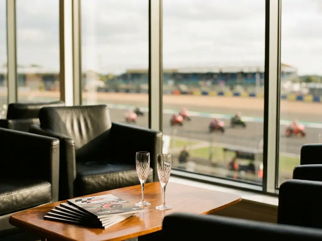 Luxury hospitality suite with leather chairs overlooking MotoGP racetrack, champagne glasses on wooden table, motorcycles racing