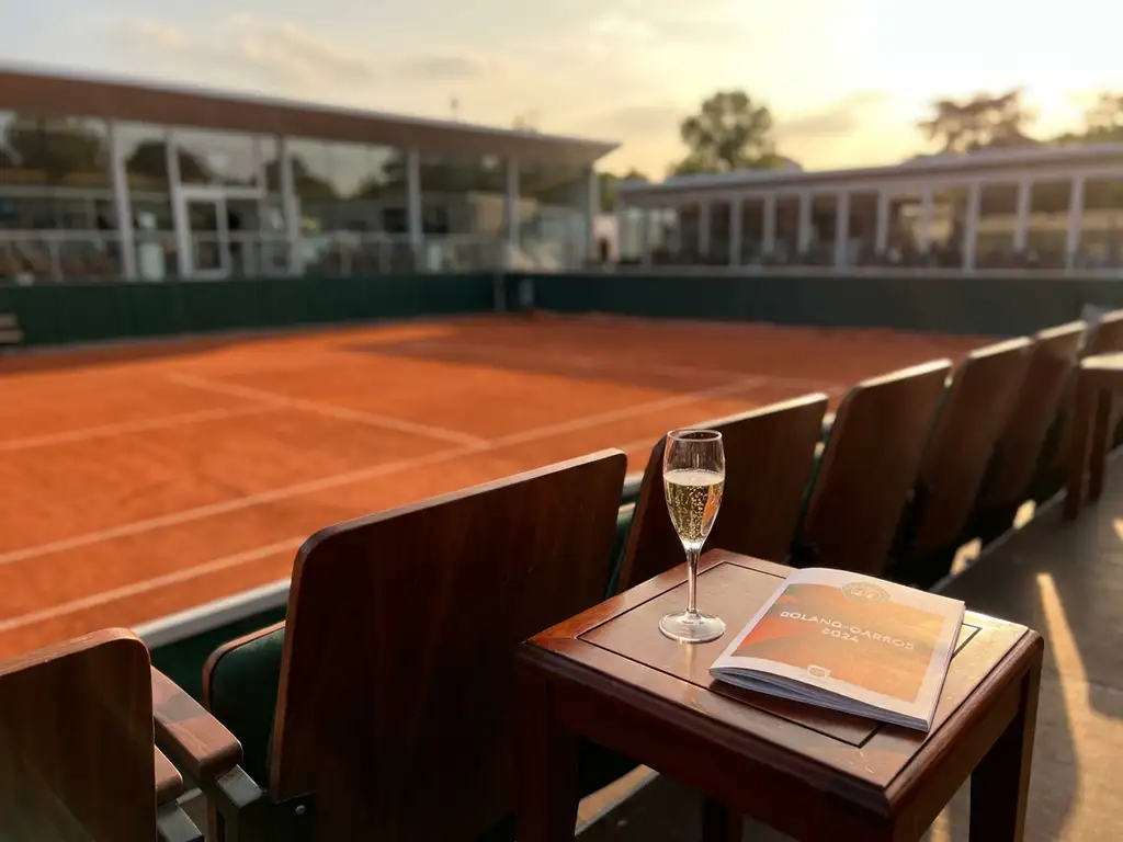 Tennis court with champagne flute and program on wooden table in foreground, corporate hospitality boxes in background